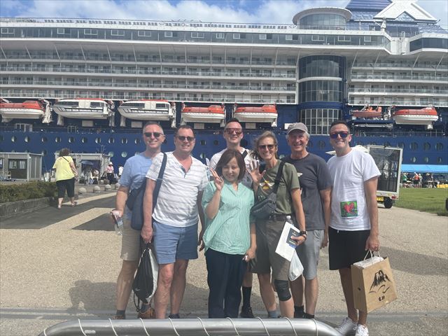 Tour guests taking a commemorative photo with their guide at the port ,cruise ship in the background. One person holds a paper bag from Takasago Sake Brewery. 
