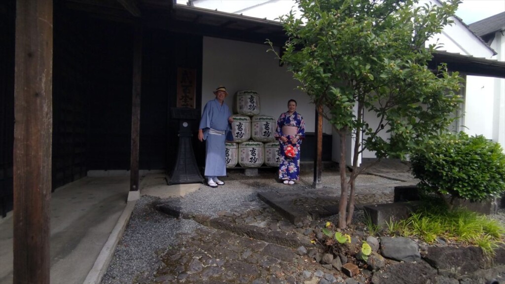 A man and a young woman in traditional Japanese kimono posing in front of sake barrels at Takasago Brewery.