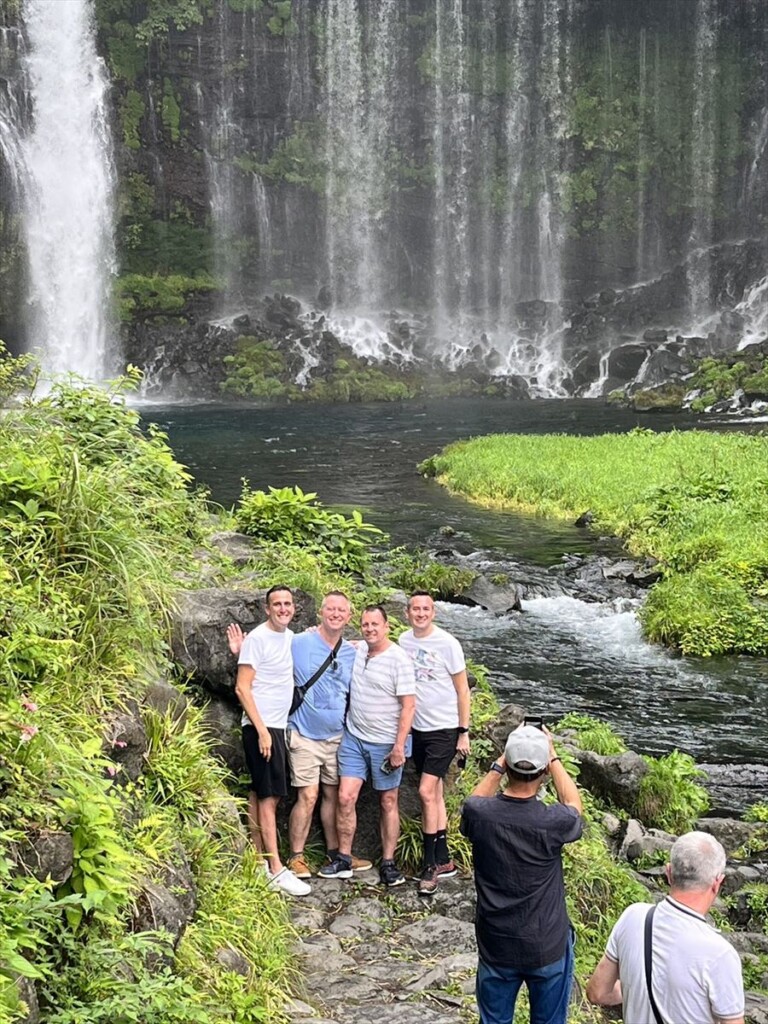 Four men smiling and posing in front of the graceful Shiraito Falls.