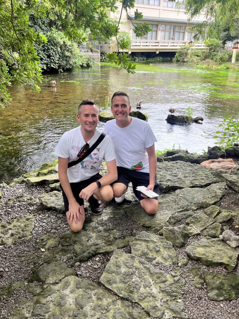 “Two men crouching and smiling beside the spring-fed Wakutama Pond.