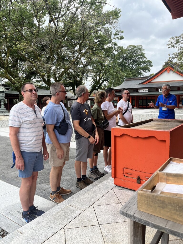 Five tourists listening to a guide near the shrine’s offertory box.