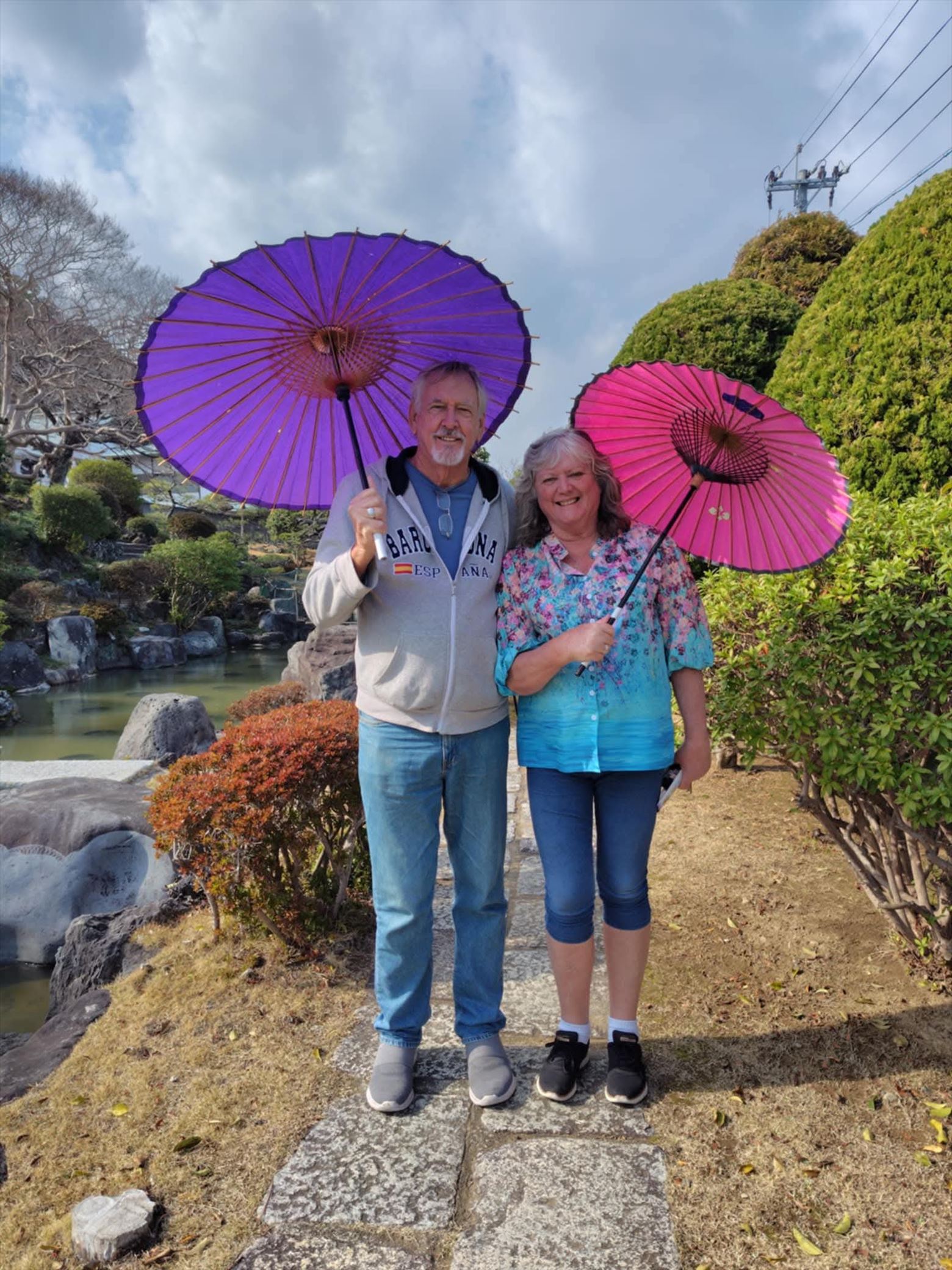 A smiling couple standing side by side in the traditional Japanese garden of Ryugeji Temple, holding purple and pink bangasa umbrellas.