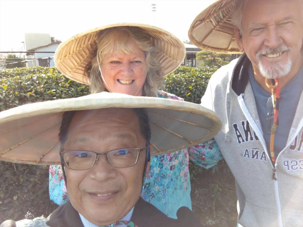Close-up of two tour guests and local driver Ted smiling together in the tea fields behind the Shizuoka Tea House, all wearing traditional straw hats (sugagasa).