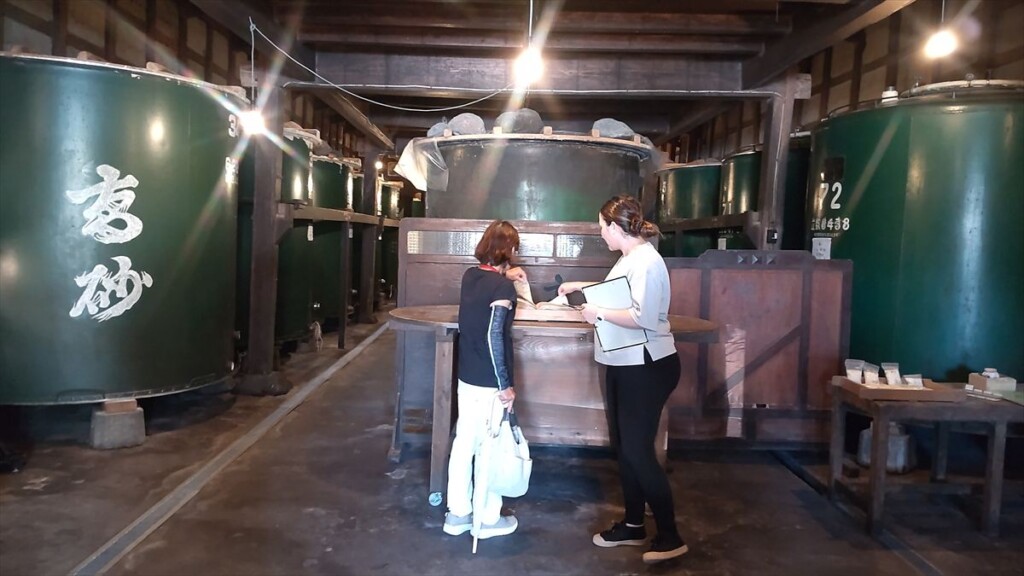 Two women reading materials in front of large fermentation tanks during a sake tour.