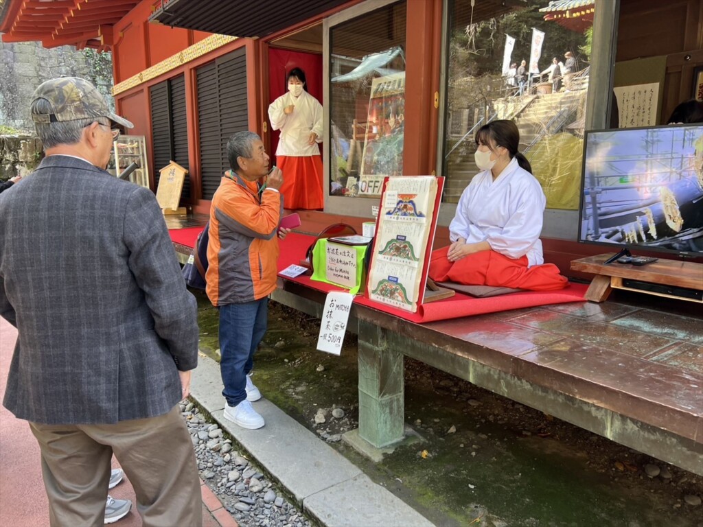 Ａ shrine maiden dressed in a traditional white kimono and red hakama is tolking with a tour guide．
