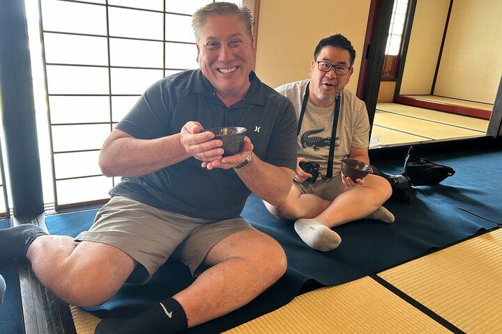 Smiling male guest holding tea after the ceremony in Shimada, with another participant in the background holding a camera, enjoying the cultural moment、
